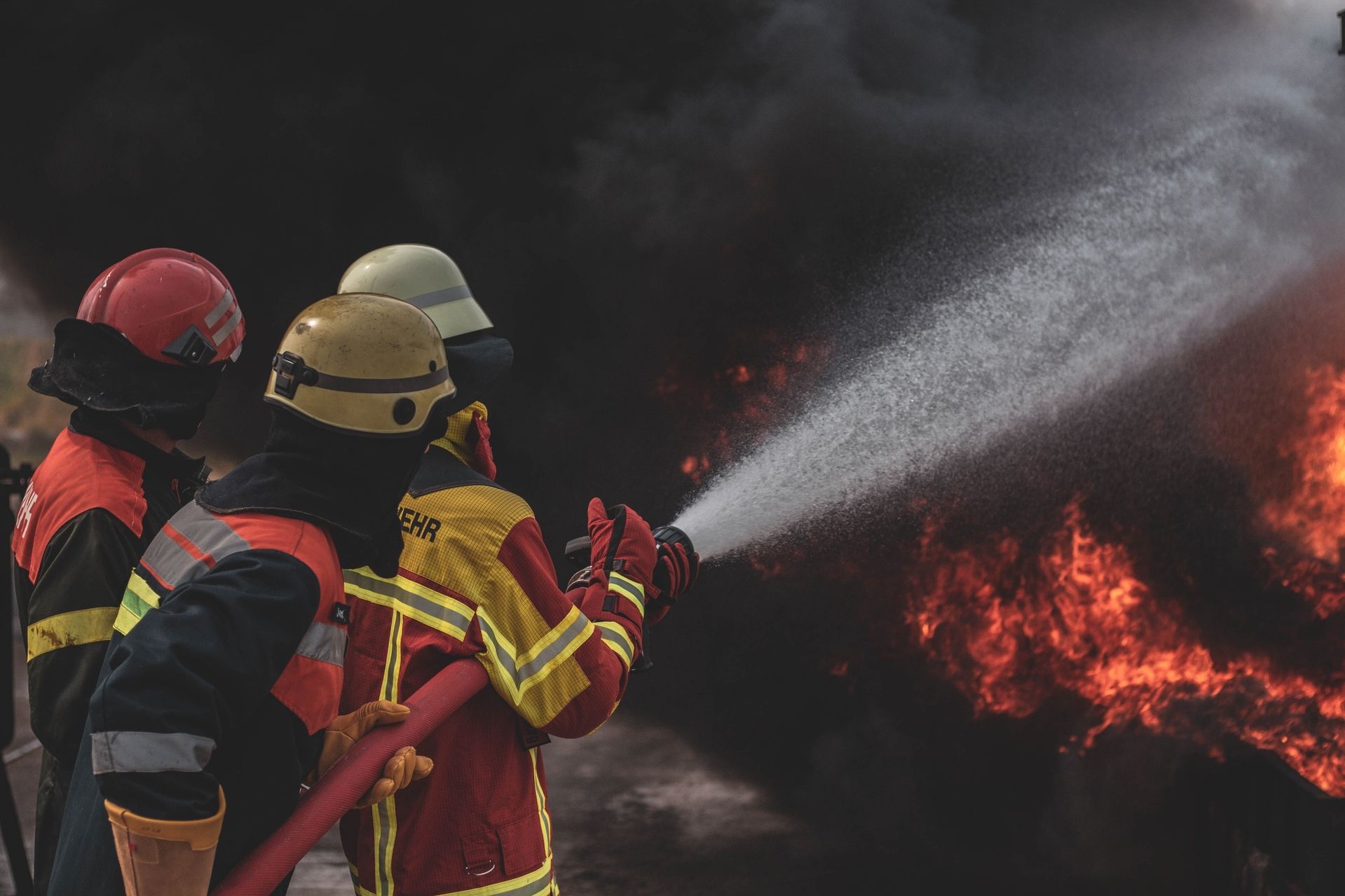 Drei Feuerwehrleute mit Helmen und Schutzkleidung löschen mit einem Schlauch ein großes Feuer